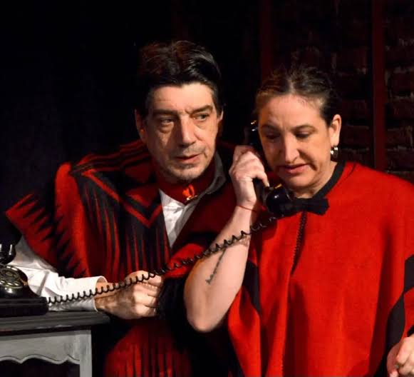 Dos actores conversan en un escenario, uno sostiene un teléfono mientras la otra escucha atentamente. Ambos visten ponchos rojos y negros.